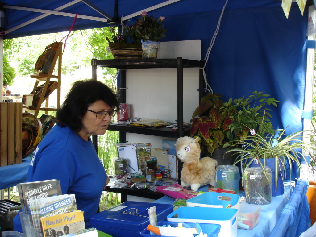 Eine Frau mit dunklem Haar sitzt an einem Stand unter einem blauen Baldachin, umgeben von Pflanzen, Büchern, kleinen Geschenkartikeln und einem ausgestopften Lama. Regale und Ausstellungsstücke sind ordentlich auf den Tischen angeordnet.