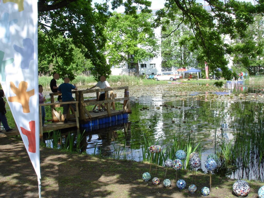 Menschen stehen auf einem kleinen Holzsteg an einem von Bäumen umgebenen Teich. Bunte, mosaikartig angeordnete Kugeln schmücken das grasbewachsene Ufer. Im Vordergrund ist eine weiße Flagge mit bunten Formen zu sehen, und im Hintergrund sind Zelte zu erkennen.