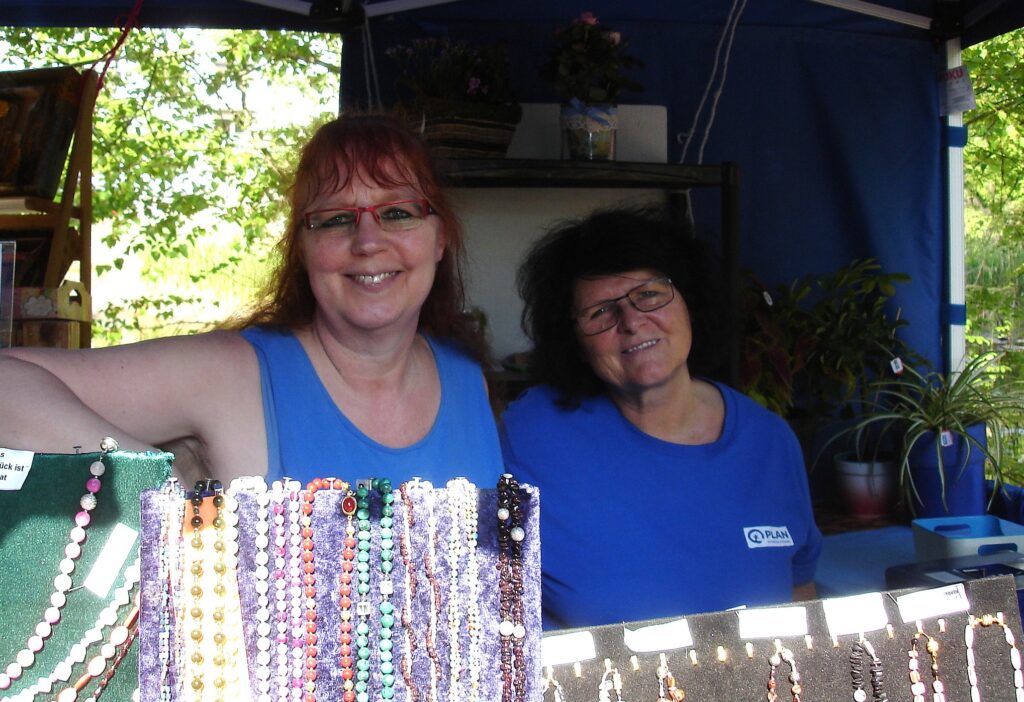 Zwei Frauen mit blauen Oberteilen stehen lächelnd hinter einer Auslage mit buntem Perlenschmuck an einem Marktstand im Freien, mit Pflanzen und verschiedenen Gegenständen im Hintergrund.