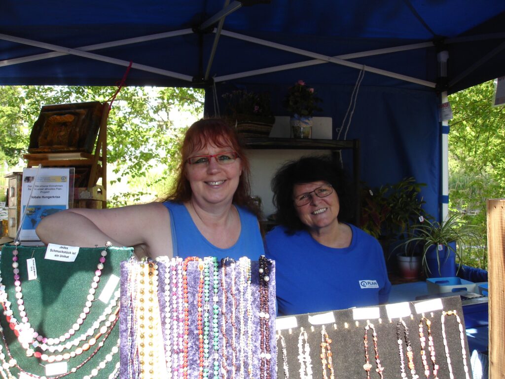 Zwei Frauen in blauen Hemden stehen hinter Tischen mit buntem Perlenschmuck an einem Marktstand im Freien und lächeln in die Kamera. Im Hintergrund sind grüne Bäume zu sehen.