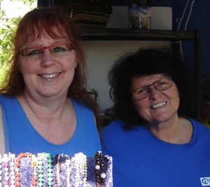 Zwei Frauen mit Brillen und blauen Oberteilen lächeln. Sie stehen hinter einer Auslage mit buntem Perlenschmuck, mit Regalen und einer Pflanze im Hintergrund.