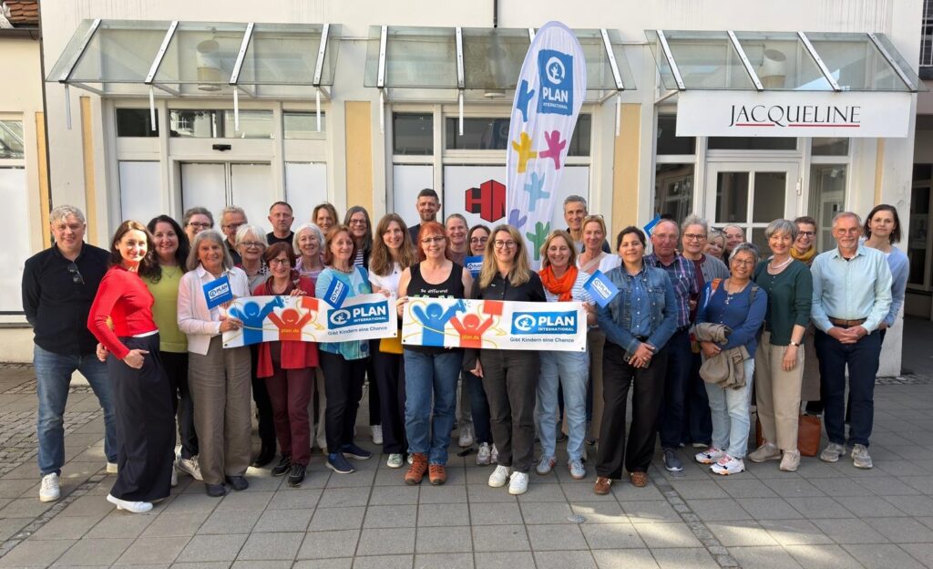 A large group of people stand smiling outdoors, holding colorful PLAN International banners and signs. They are in front of a building with a sign reading Jacqueline and a tall PLAN flag, suggesting a group event or gathering.