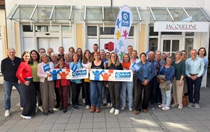 A group of about 35 people are standing outside a building, smiling at the camera. Some hold banners and a flag featuring the Plan International logo. The building has large windows and a sign reading JACQUELINE.