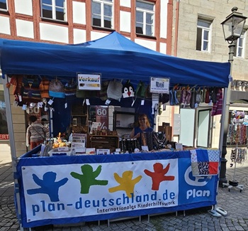 Ein blauer Zeltstand der Organisation Plan Deutschland, an dem auf einem Stadtplatz gestrickte Kindermützen und Accessoires verkauft werden.