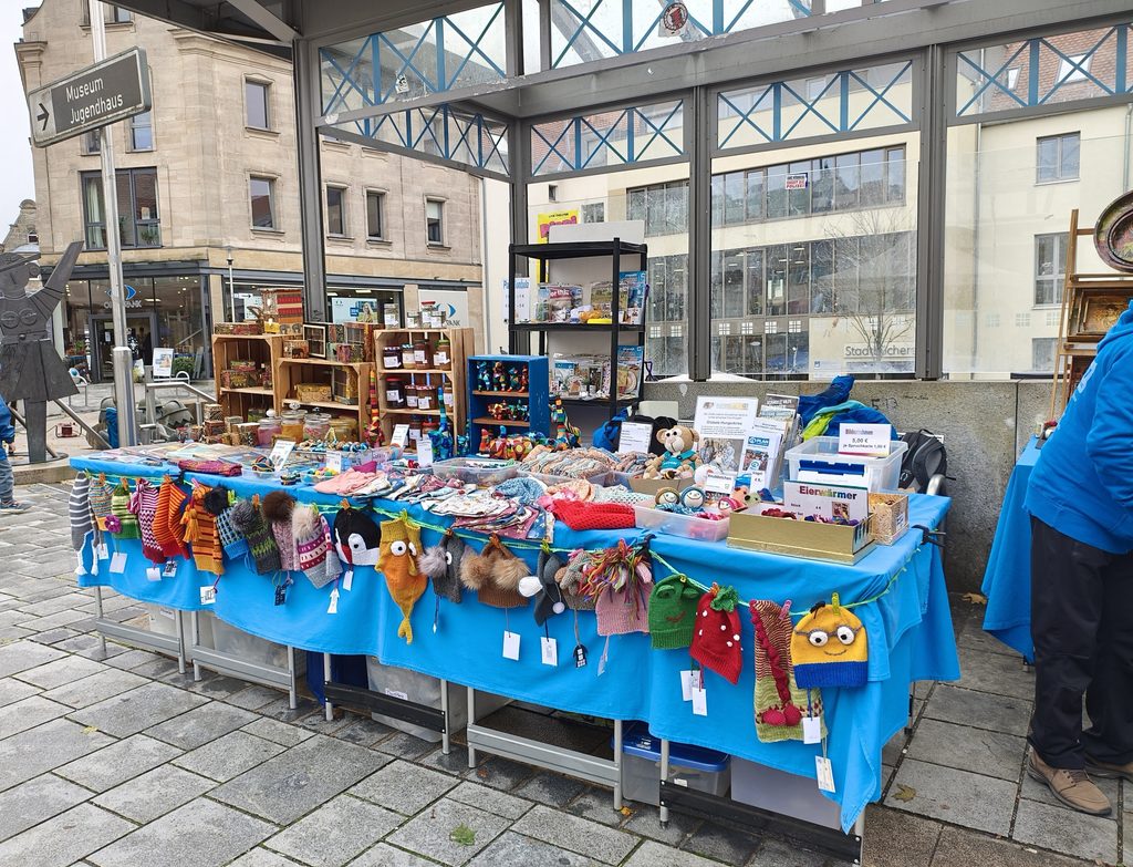 Ein Marktstand unter einem Glasvordach, an dem verschiedene handgestrickte Artikel verkauft werden, darunter Kindermützen mit Tiermotiven, die auf einem langen Tisch mit blauer Tischdecke ausgestellt sind.