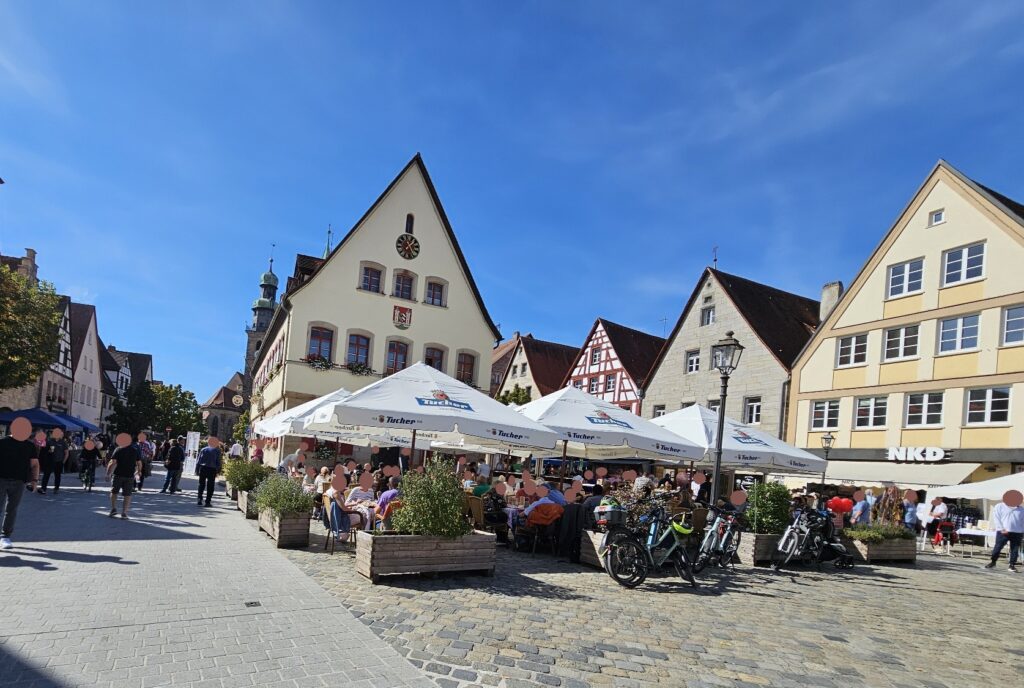Menschen, die an Tischen im Freien unter weißen Sonnenschirmen auf einem sonnigen Stadtplatz speisen, der von historischen europäischen Gebäuden umgeben ist.