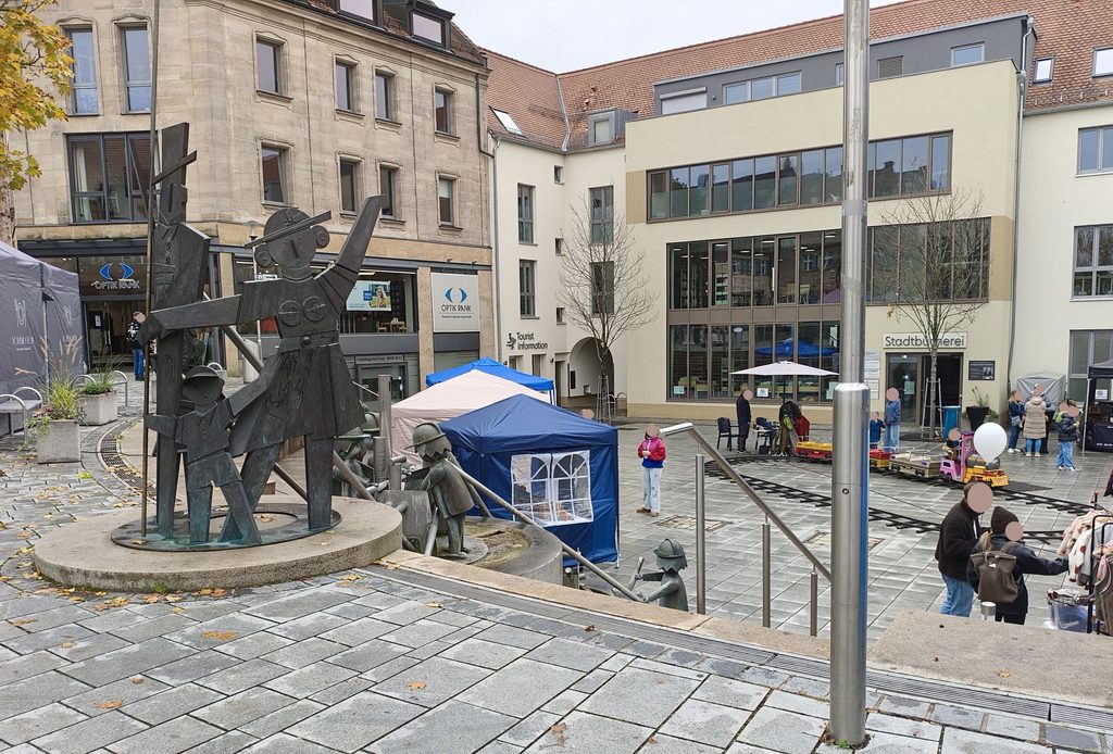 Auf einem Steinplatz vor den städtischen Gebäuden steht eine Bronzeskulptur einer Frau mit einem Kind; in der Nähe befinden sich blaue Marktstände und eine kleine Bahn für Kinder.