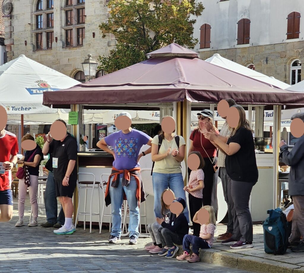 Eine Gruppe von Menschen, die vor einem Getränkestand auf einem mit Kopfsteinpflaster gepflasterten Stadtplatz stehen und sitzen.