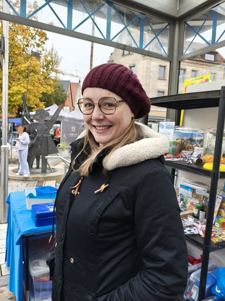 Eine Frau mit Brille und einer kastanienbraunen Baskenmütze steht an einem Marktstand im Freien und lächelt.