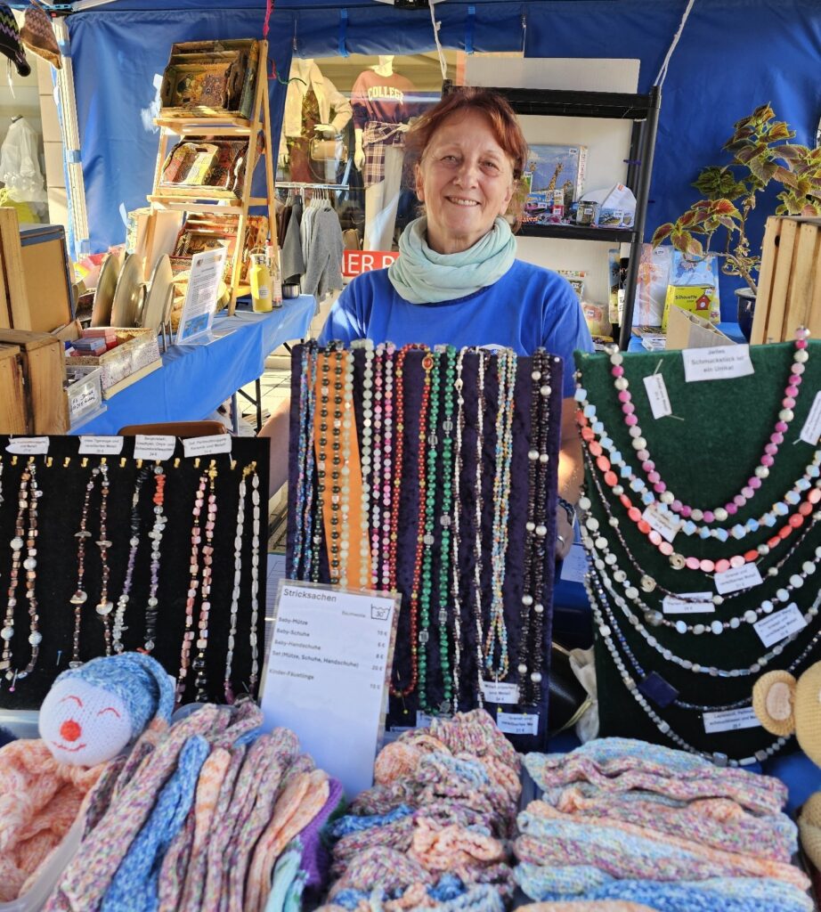 Hinter einem Marktstand, auf dem handgefertigte Perlenketten und Strickwaren mit Preisschildern ausgestellt sind, steht eine lächelnde Frau.