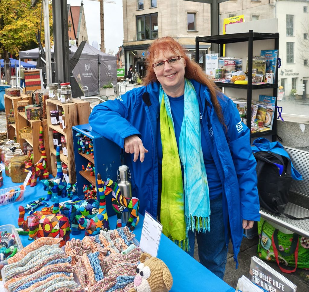 Hinter einem Marktstand, auf dem bunte Tierfiguren und Strickwaren ausgestellt sind, steht eine Frau mit rötlichem Haar und einer blauen Jacke.