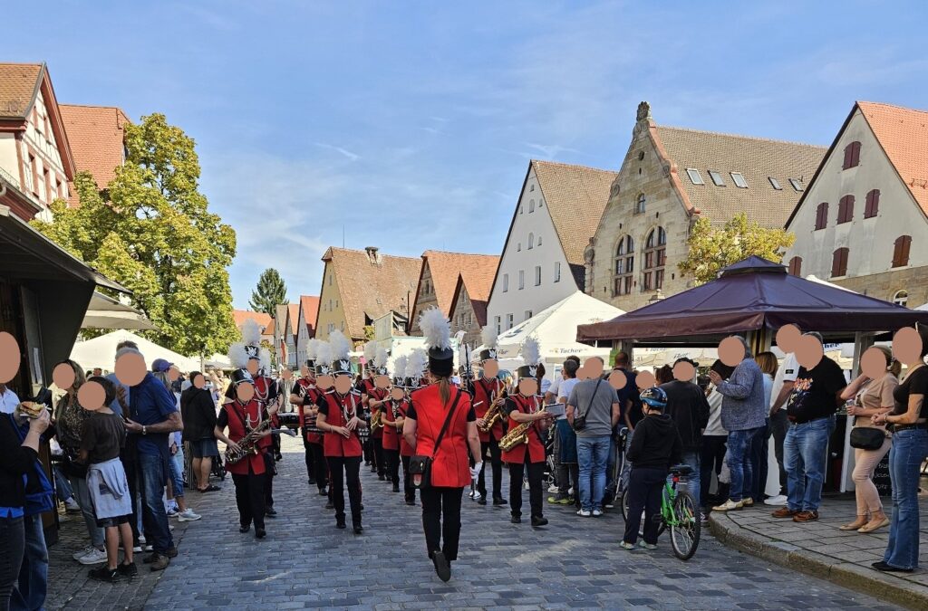 Eine Blaskapelle in roten Uniformen und mit Federhüten tritt auf einer Kopfsteinpflasterstraße auf einem von historischen Gebäuden gesäumten Stadtplatz auf.