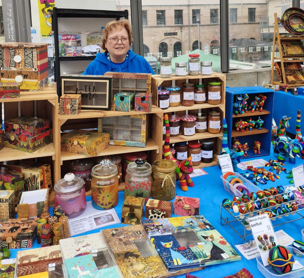 Eine Frau steht hinter einem Marktstand, der mit handgefertigten Artikeln gefüllt ist, darunter dekorative Holzschachteln, Gläser mit Konfitüre und bunte kleine Skulpturen.