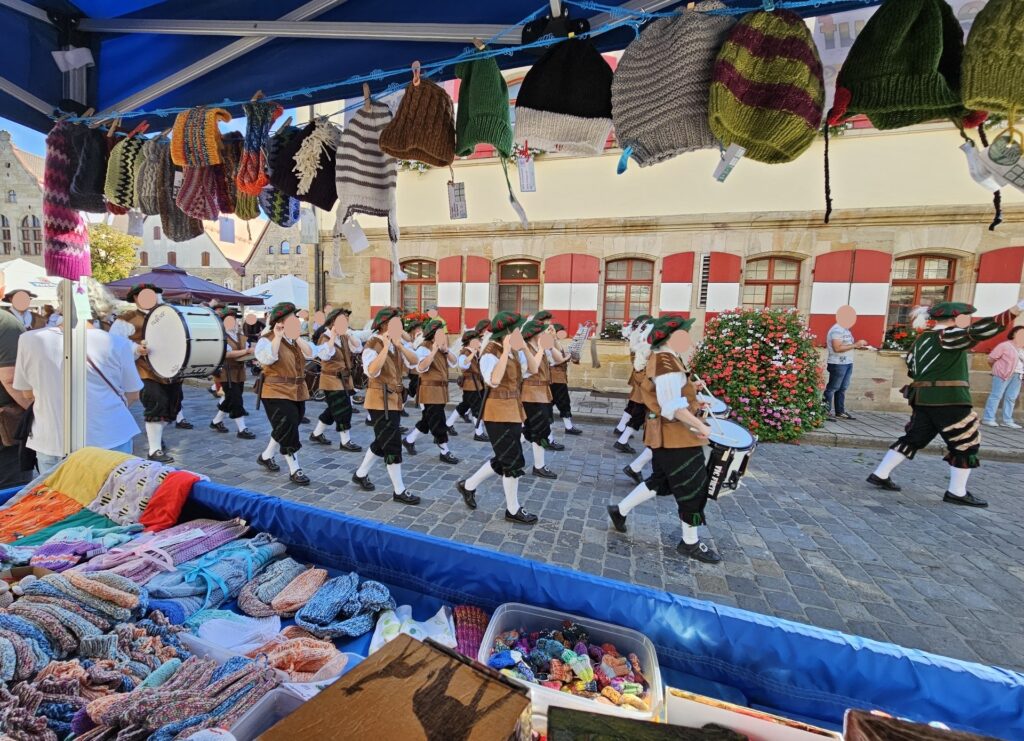 Eine Blaskapelle in traditioneller deutscher Tracht spielt auf einer Kopfsteinpflasterstraße, im Hintergrund ist ein Marktstand mit gestrickten Mützen und Accessoires zu sehen.