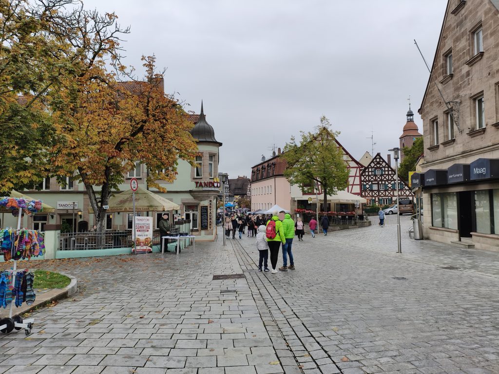 Menschen, die an einem bewölkten Tag über einen mit Kopfsteinpflaster gepflasterten Stadtplatz spazieren, der von Fachwerkhäusern und Geschäften gesäumt ist.