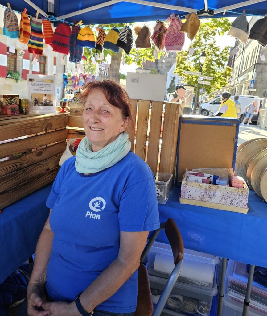 Eine Frau, die ein blaues T-Shirt von Plan International und einen hellblauen Schal trägt, sitzt an einem Stand voller handgestrickter Mützen.