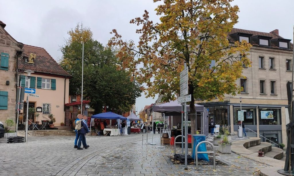 Menschen spazieren über einen mit Kopfsteinpflaster gepflasterten Stadtplatz mit Marktständen und herbstlich gefärbten Bäumen.