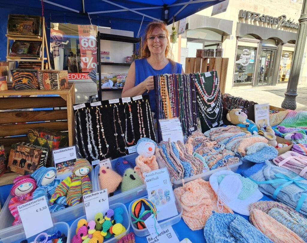 Eine Frau steht hinter einem Marktstand, auf dem handgefertigte Perlenketten, gehäkelte Stofftiere und gestrickte Babykleidung ausgestellt sind.