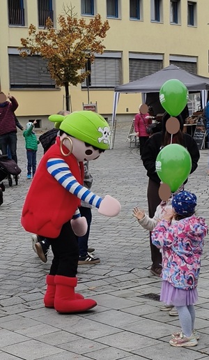 Ein als Pirat verkleidetes Maskottchen unterhält sich auf einem gepflasterten Platz mit einem kleinen Kind, das grüne Luftballons in der Hand hält.