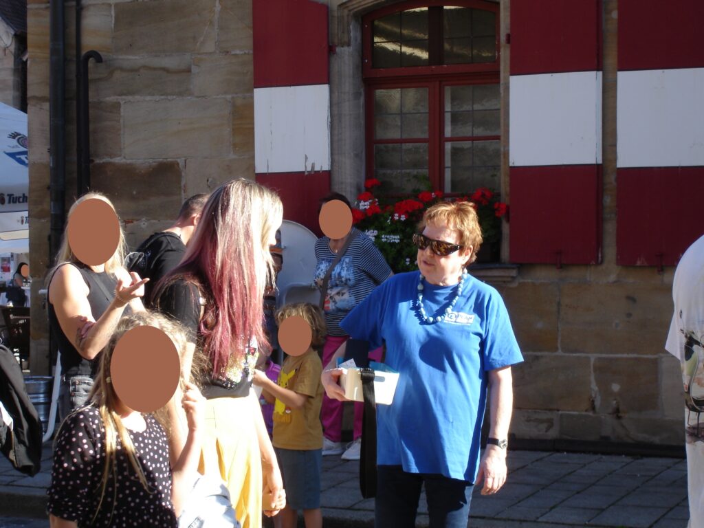 Eine Frau in einem blauen T-Shirt und mit Sonnenbrille hält eine weiße Schachtel in der Hand, während sie sich im Freien vor einem Steingebäude mit einer Gruppe von Menschen unterhält.