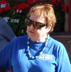Eine Frau mit kurzen braunen Haaren, die eine Sonnenbrille und ein blaues T-Shirt trägt und eine Perlenkette um den Hals hat.