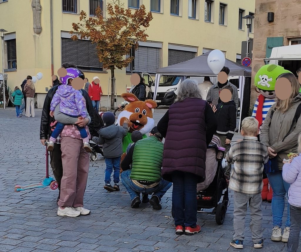 Auf einem mit Kopfsteinpflaster gepflasterten Stadtplatz spielen Menschen und Kinder mit einem braunen Kaninchen-Maskottchen und einem grünen Maskottchen mit Totenkopfhut.