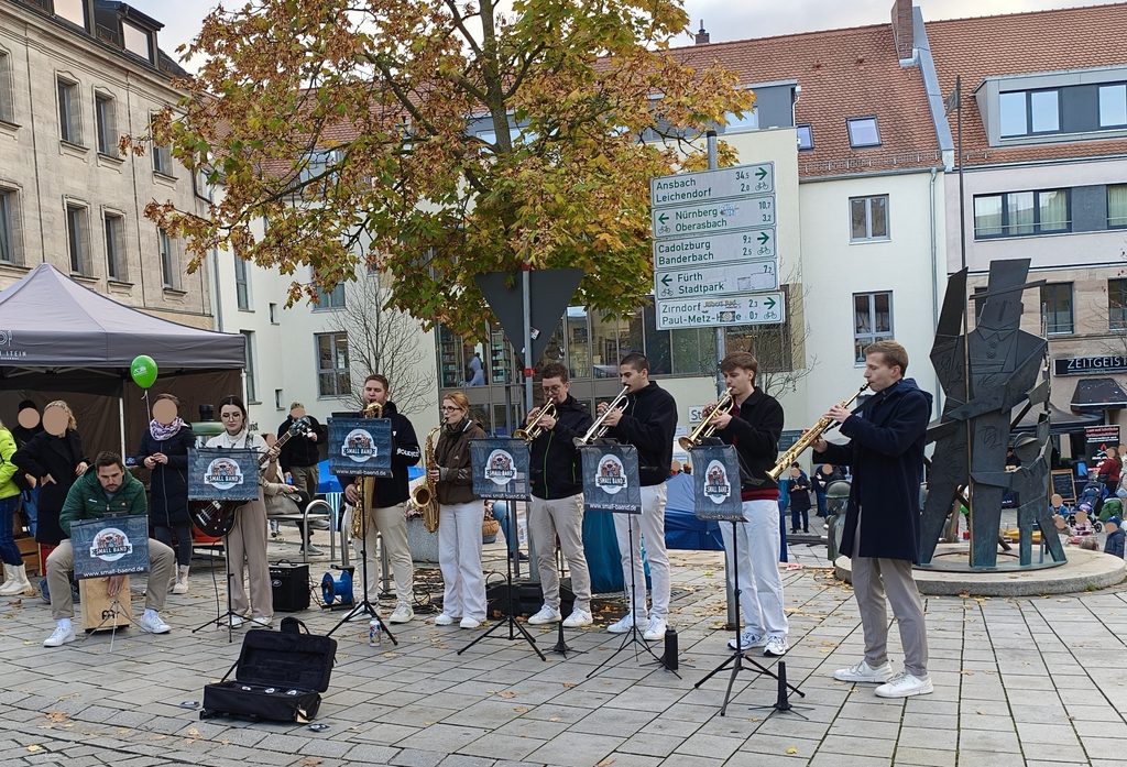 Eine Gruppe von Musikern, die auf einem Platz im Freien Blechblasinstrumente und Saxophon spielen, im Hintergrund ein Wegweiser, der auf deutsche Städte in der Umgebung hinweist.