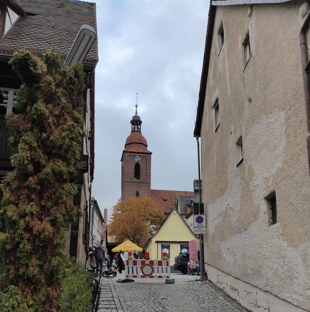 Eine Kopfsteinpflasterstraße führt auf einen Kirchturm zu, der durch eine rot-weiße Bauabsperrung in der Nähe eines gelben Gebäudes versperrt ist.