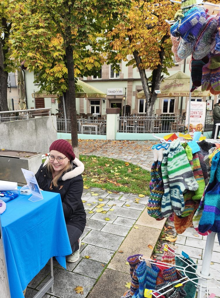 Eine Frau mit einer kastanienbraunen Mütze und einer Brille sitzt an einem blau gedeckten Tisch neben einem Verkaufsständer mit bunten, handgestrickten Socken auf einem Markt im Freien.