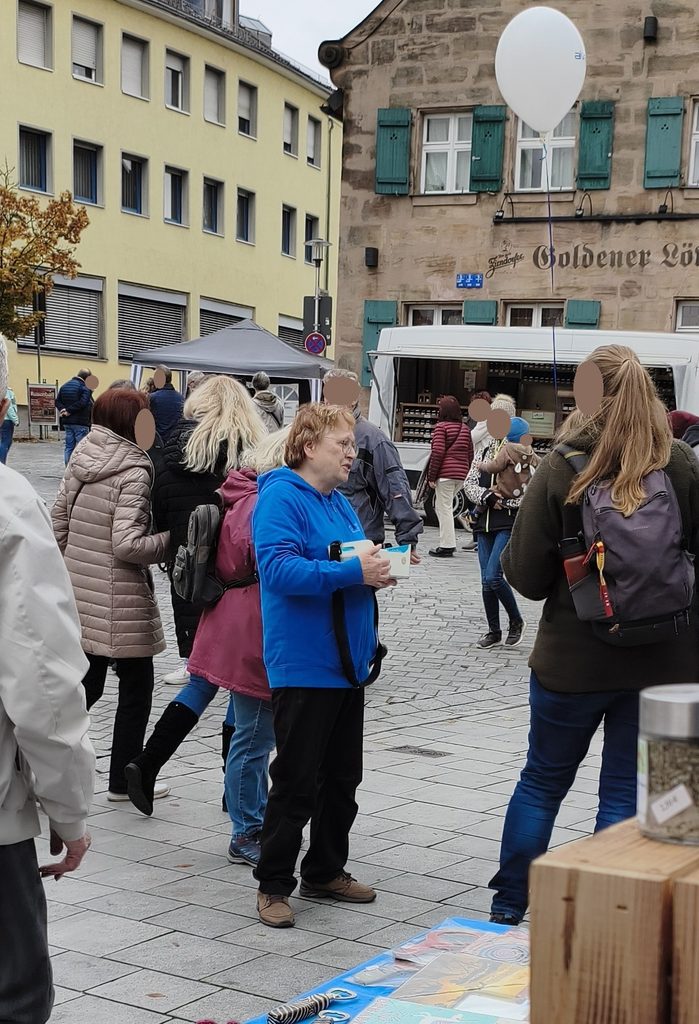 Menschen, die über einen mit Kopfsteinpflaster gepflasterten Stadtplatz gehen, auf dem ein Imbisswagen und ein weißer Ballon zu sehen sind.
