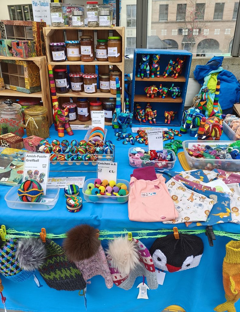 Eine Marktstandauslage mit Gläsern voller Konfitüren, handgefertigten Tierfiguren, Strickwaren und buntem Spielzeug, versehen mit Preisschildern in deutscher Sprache.
