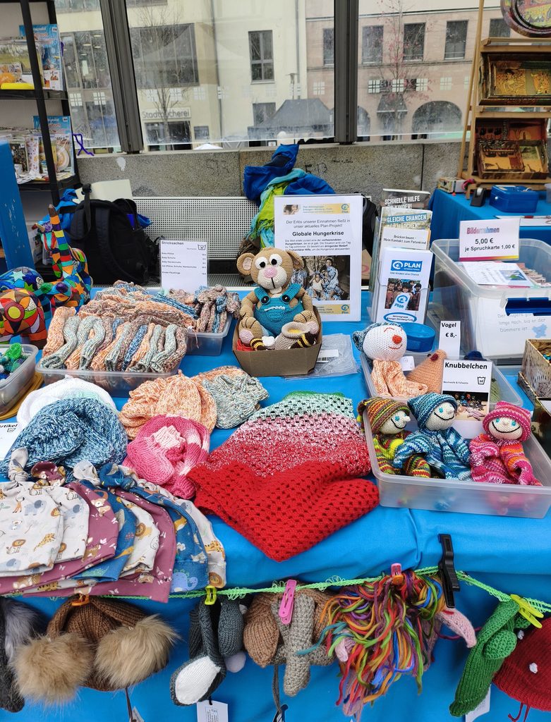 Ein Tisch auf einem Kunsthandwerksmarkt, bedeckt mit handgefertigter Babykleidung, gestrickten Mützen und gehäkelten Puppen, mit Informationsschildern zu einem Spendenprojekt von Plan International.