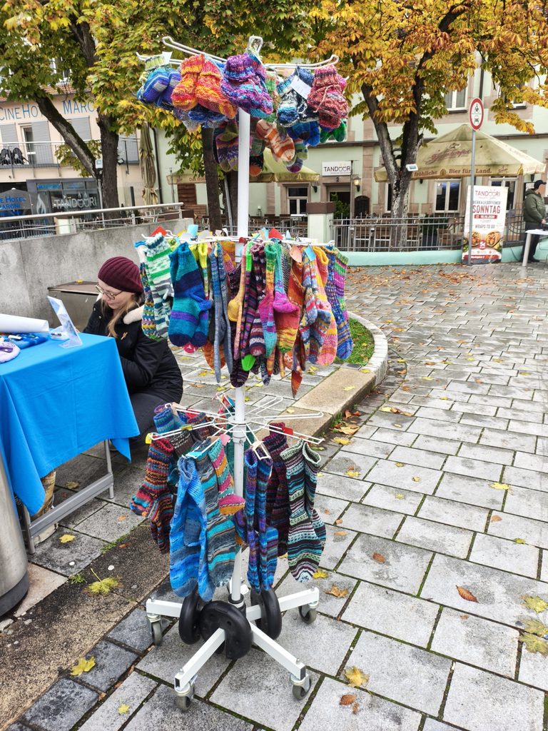 Ein drehbarer Ausstellungsständer, gefüllt mit bunten, handgestrickten Socken, an einem Marktstand im Freien.