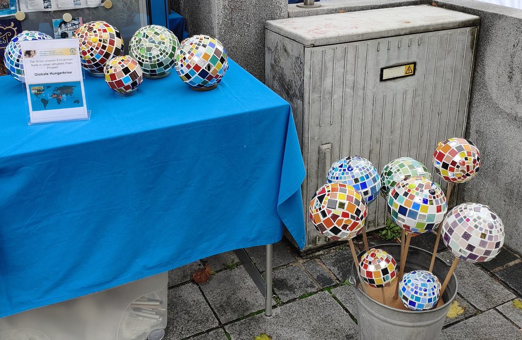 Dekorative Mosaikgloben, die auf einem blau bezogenen Tisch ausgestellt sind und in einem Metalleimer stehen.