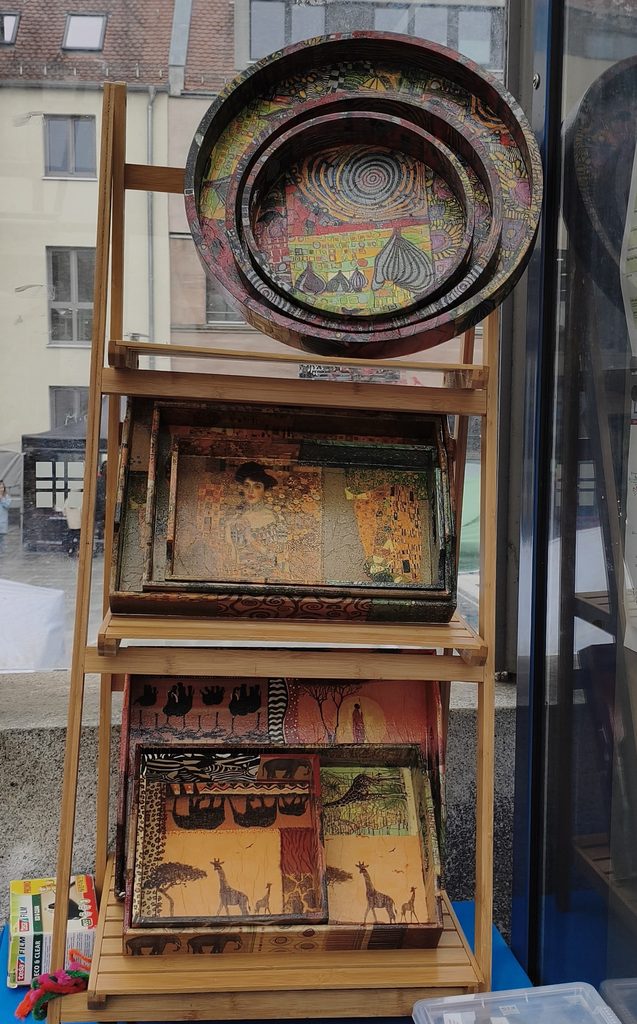 Ein mehrstufiger Ausstellungsständer aus Holz, auf dem dekorative Tabletts mit kunstvollen Mustern präsentiert werden, darunter ein Porträt, das an ein Werk von Gustav Klimt erinnert, sowie Tiersilhouetten.