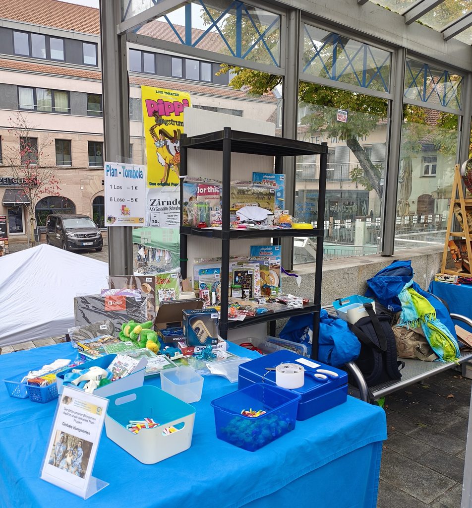 Ein Tombola-Stand auf einem Markt im Freien mit einem Plakat für eine Plan-Tombola und einem schwarzen Regal, das mit Preisen und Spielzeug gefüllt ist und auf einer blauen Tischdecke steht.