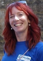 Eine lächelnde Frau mit roten Haaren, die ein blaues T-Shirt mit einem Plan-Logo trägt.
